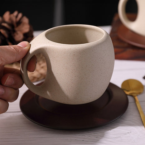 Beige Keramiktasse mit Henkel auf dunklem Holzuntersetzer, Hand hält die Tasse