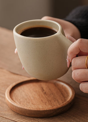 Steingut-Kaffeetasse mit Holzuntersetzer, Hand hält die Tasse am geschwungenen Griff