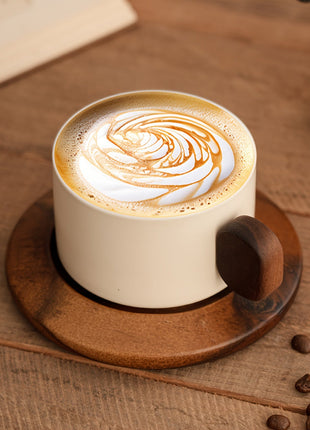 Cremefarbene Keramiktasse mit Untertasse, brauner Griff, geschäumter Latte mit Wirbelmuster.