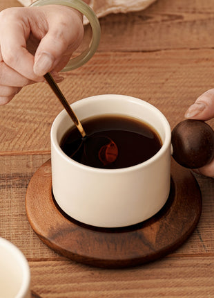 Cremefarbene Keramiktasse auf brauner Untertasse, Hand rührt Kaffee mit Löffel