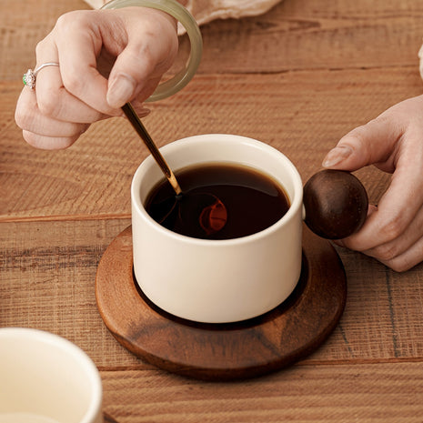 Cremefarbene Keramiktasse auf brauner Untertasse, Hand rührt Kaffee mit Löffel