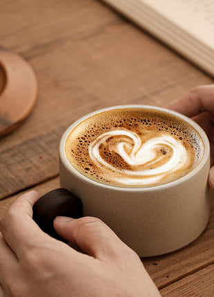Keramik-Tasse mit Untertasse, beiger Becher mit braunem Griff und handgehaltenem Cappuccino