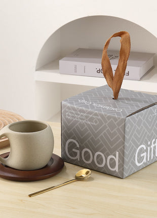 Steingut-Kaffeetasse mit braunem Holzuntersetzer neben Geschenkbox und goldenem Löffel