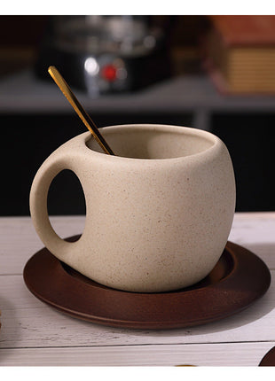 Hellbeige Steingut-Kaffeetasse auf braunem Holzuntersetzer, goldener Löffel daneben