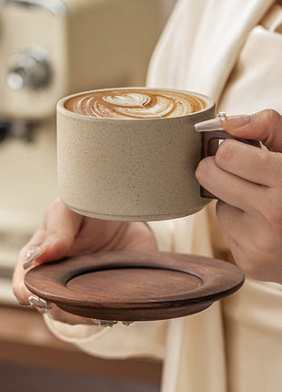 Beige Keramiktasse mit braunem Kaffee auf Holzuntersetzer, Hand hält den Henkel