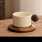 Cremefarbene Keramiktasse mit braunem Holzgriff auf Untertasse, seitlicher Blick