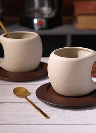 Beige Keramiktasse mit Henkel auf dunklem Holzuntersetzer, goldfarbener Löffel