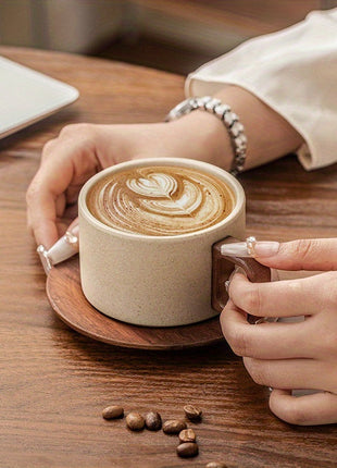 Beige Keramiktasse auf Holzuntersetzer mit Latte-Art, Hände greifen den Henkel