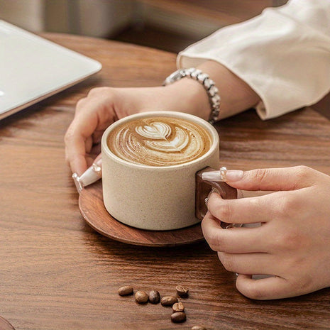 Beige Keramiktasse auf Holzuntersetzer mit Latte-Art, Hände greifen den Henkel