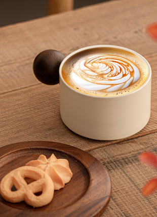 Cremefarbene Keramiktasse mit Untertasse, brauner Holzgriff, Latte mit Wirbelmuster.
