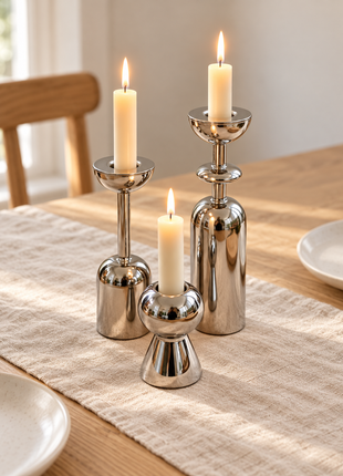 Three silver-tone geometric candle holders with ivory candles on a linen runner and light oak dining table.